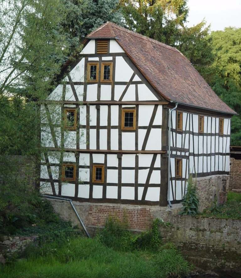 Fachwerkhaus mit braunen Holzrahmen und Holzfenstern, umgeben von Grün.