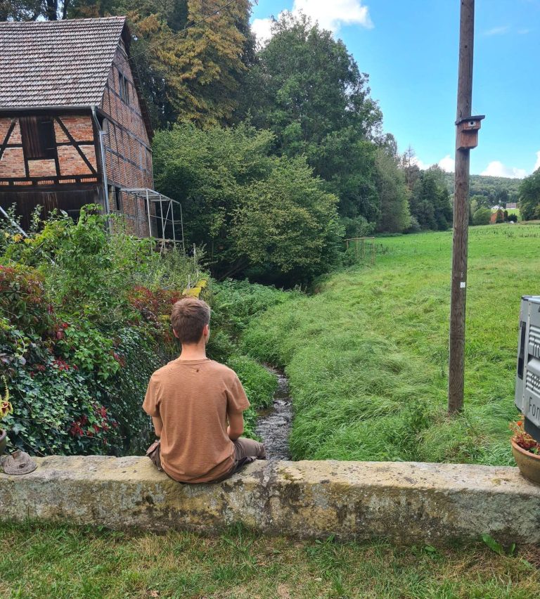 Junger Mann sitzt auf einer Mauer neben einem Bach, umgeben von grüner Landschaft.