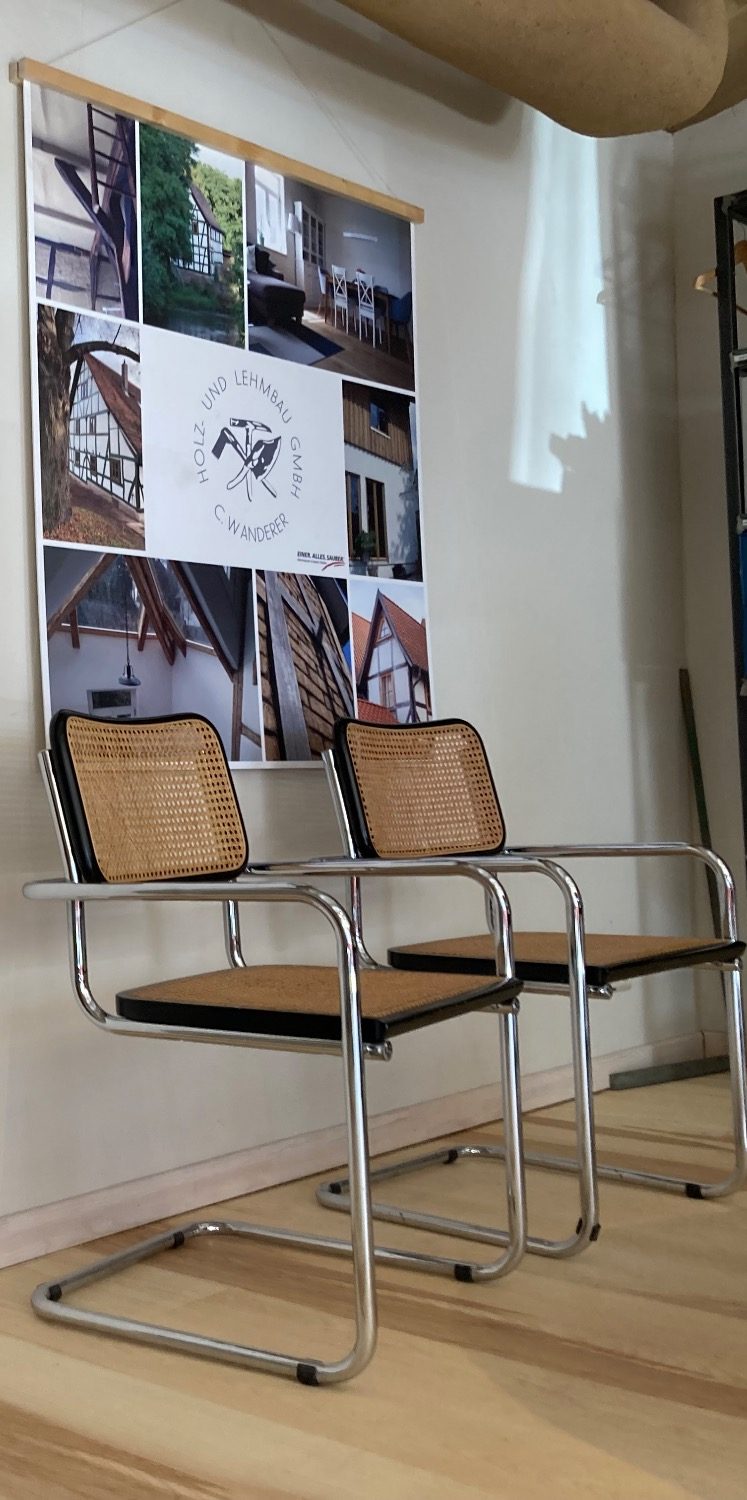 Zwei metallene Stühle (Modell Cesca von Thonet) mit geflochtener Sitzfläche vor einer Bilderwand.