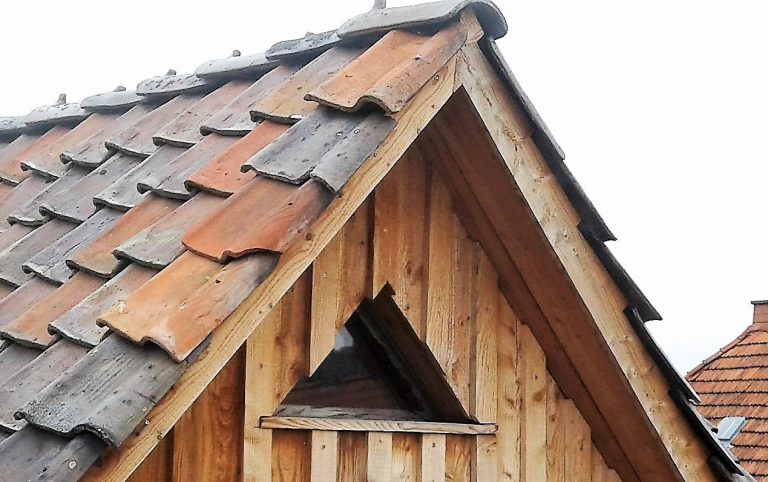 Dach eines Fachwerkhauses mit Lärchendeckelschalung, historischen Dachziegeln und dreieckigem Fenster oben.